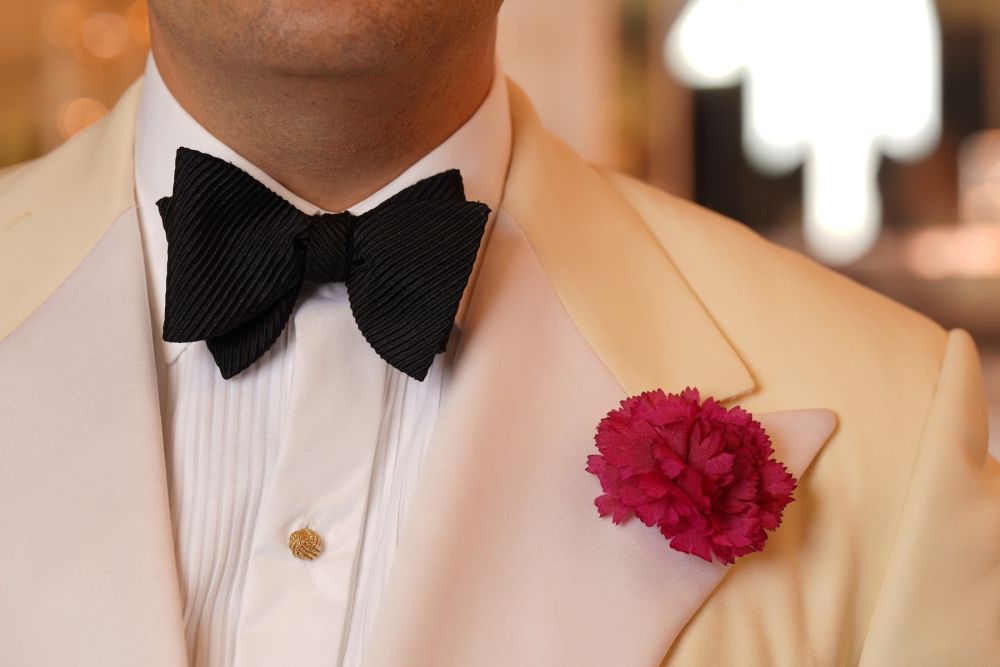 Red Mini Carnation Silk Boutonniere combine with a black grosgrain bow tie, gold shirt studs, white dress shirt and off-white tuxedo  jacket