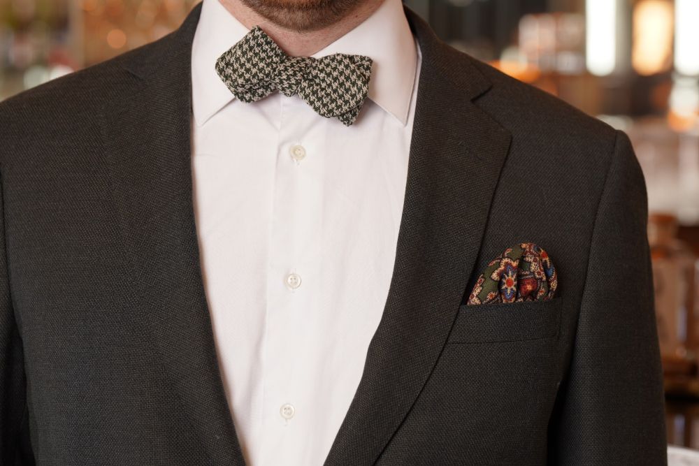 Moss Green Silk Wool Pocket Square with Printed geometric medallions in blue, red, black with eggshell contrast edge combined with a Houndstooth Silk Bourette Bow Tie Dark Green Pearl Grey in a dark gray suit