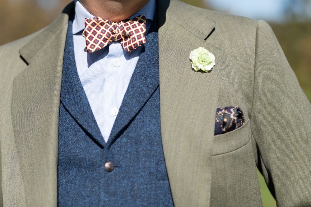 Ancient Madder Silk Bow Tie in Yellow, Red Blue and Orange Diamond Pattern in light blue & white striped long sleeves, blue vest and light brown suit with pale green carnation boutonniere and navy blue silk wool pocket square - all accessories by Fort Bel