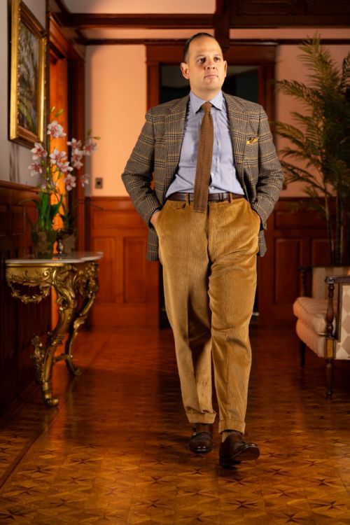 Raphael wearing a checked brown and yellow jacket, checked light blue and yellow shirt, brown knit tie, paired with the camel corduroy trousers. 
