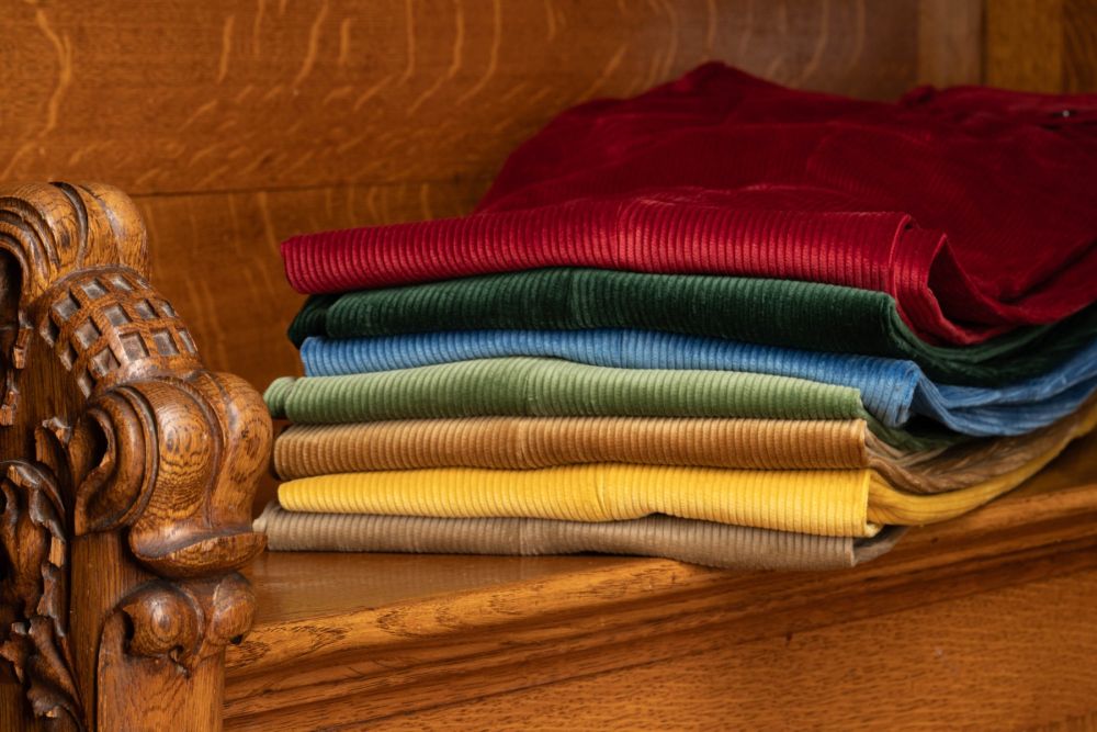 Garnet Red, British Racing Green, Azure Blue, Sage Green, Camel, Goldenrod Yellow and Pale Taupe Corduroy Trousers, Model Stancliffe by Fort Belvedere neatly stacked on a wooden bench.