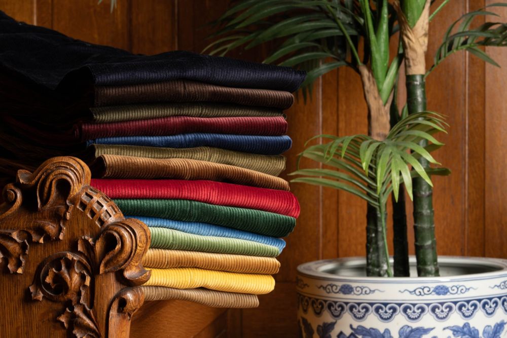 All 14 colors of Corduroy Trousers, Model Stancliffe by Fort Belvedere neatly stacked on top of each other on a wooden hand carved bench next to a potted palm tree