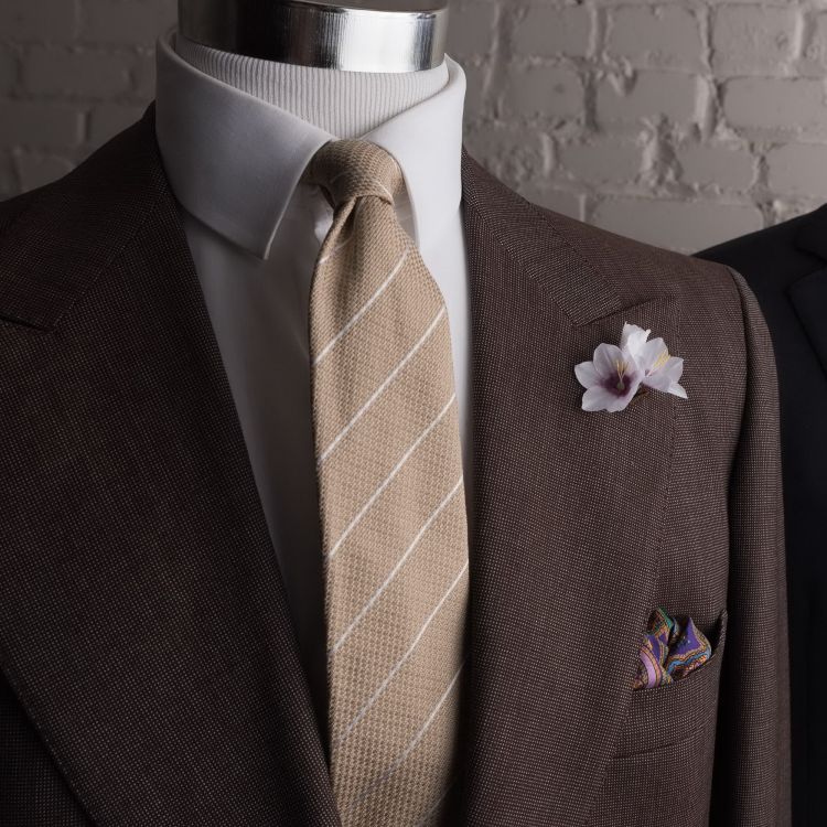 White Ixia boutonniere flower with purple paisley pocket square and beige 3 fold necktie By Fort Belvedere